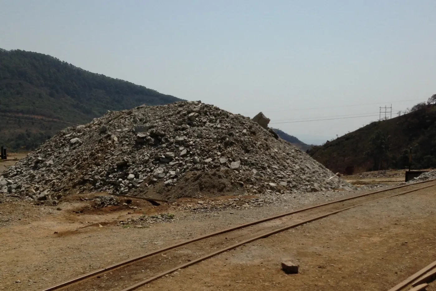 A stockpile of broken rock beside narrow-gauge rail tracks, with wooded hills behind